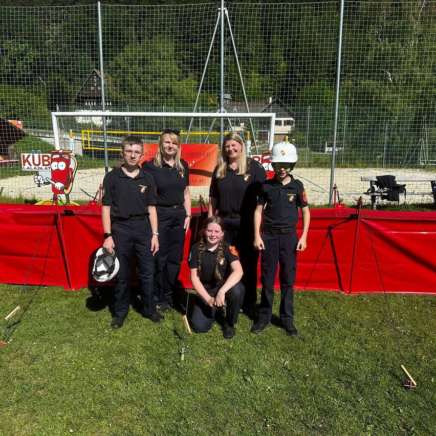 Fünf Personen, darunter zwei Frauen und drei Männer, stehen auf einem Rasenfeld und tragen Uniformen. Eine Frau hält einen Helm. Ein roter Zaun steht hinter ihnen.