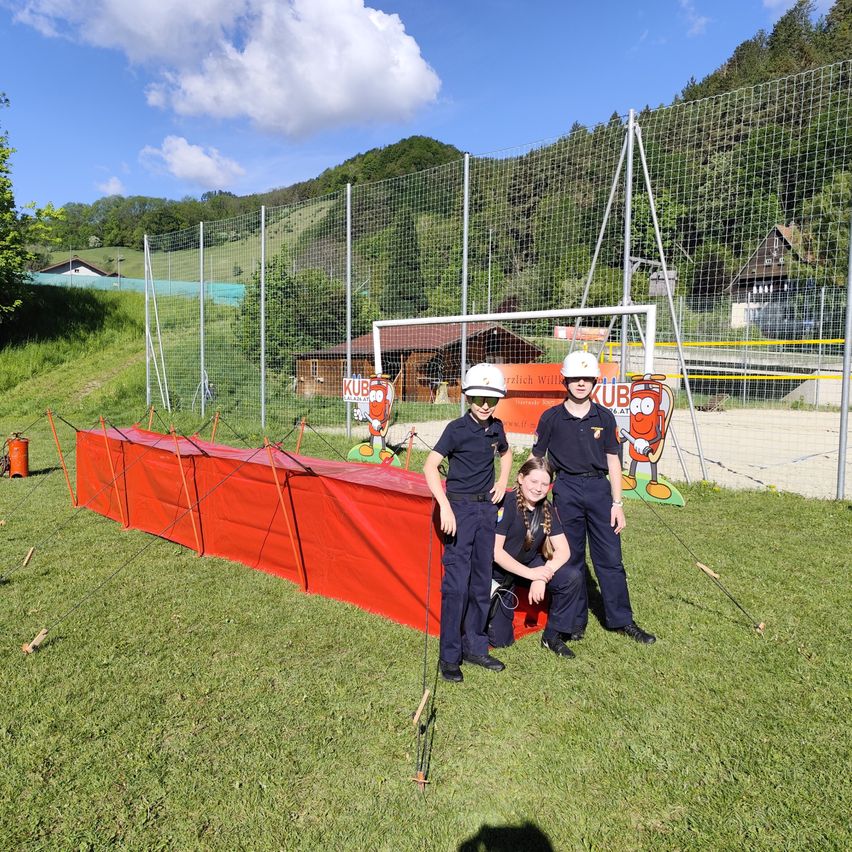 Drei Personen in Uniformen stehen neben einer roten Barriere. Sie posieren für ein Foto. Dahinter befindet sich ein Metallzaun und ein Haus. In der Ferne sind Bäume und Berge zu sehen.