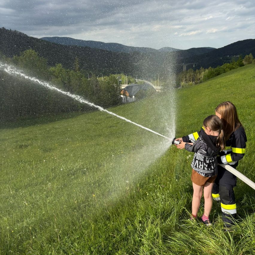 Zwei Personen stehen in einem Grasfeld und halten einen Feuerwehrschlauch. Eine spritzt Wasser, während die andere zuschaut.