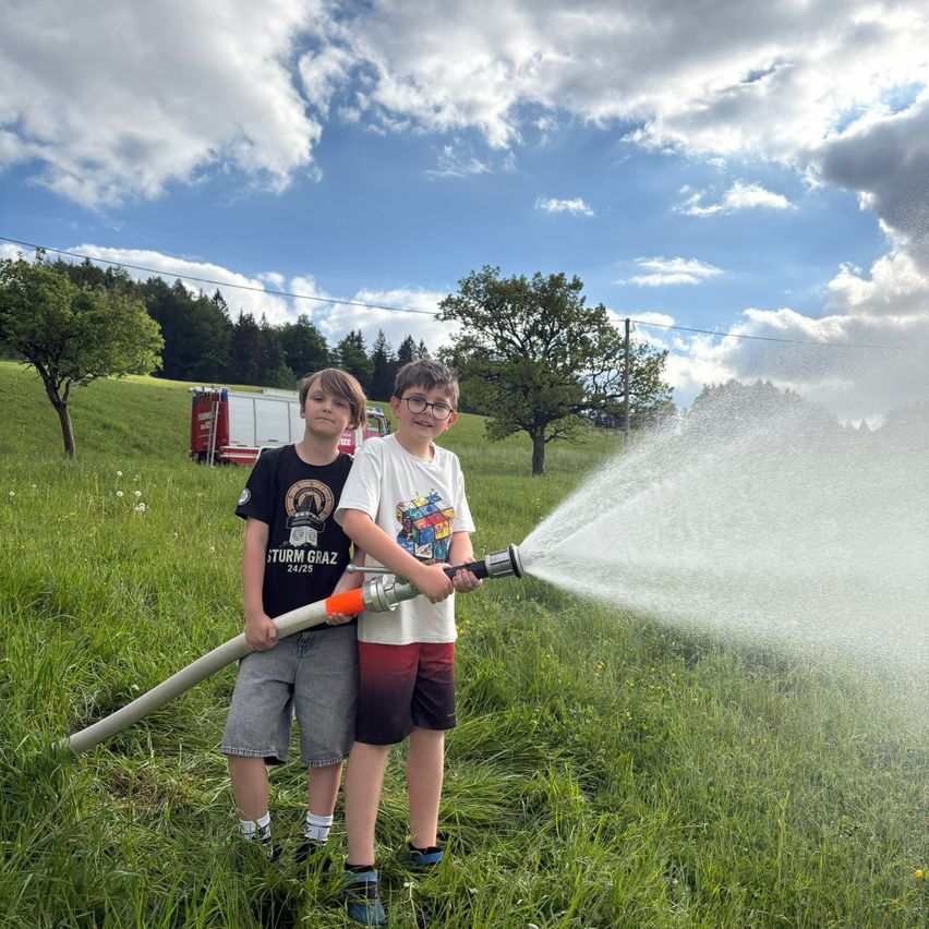 Zwei Jungen stehen in einem Grasfeld und halten einen Feuerwehrschlauch in der Hand. Im Hintergrund ist ein Feuerwehrwagen neben Bäumen geparkt. Der Himmel ist bewölkt.