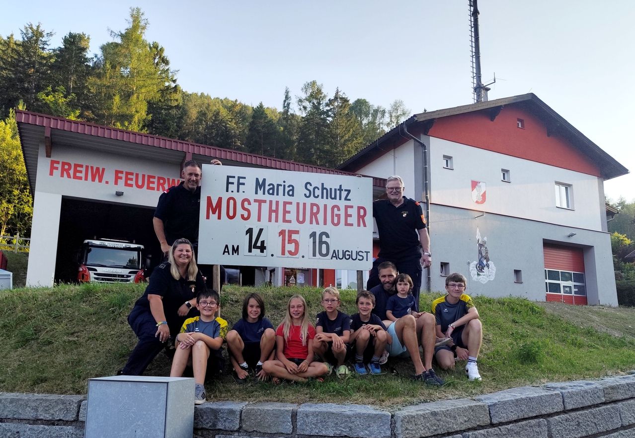 Eine Gruppe von Feuerwehrleuten und Kindern posiert für ein Foto vor einer Feuerwache mit einem Schild, das 'F.F. Maria Schutz Mostheueriger 14.15.16 August' steht.