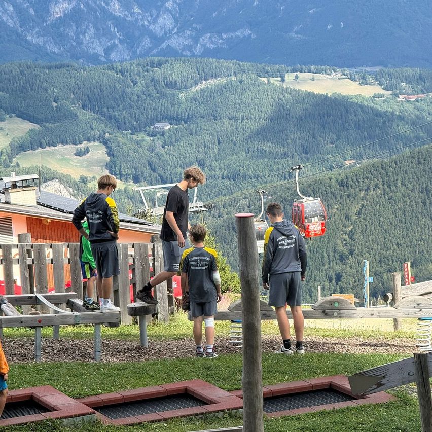Five boys stand on a wooden platform in a mountainous area, with a cable car in the background.