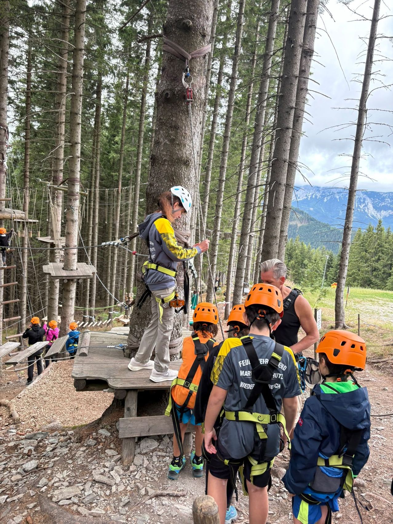 A group of people in safety gear are gathered around a tree in a forest. A woman is standing on a wooden platform, possibly giving instructions. The children are wearing orange helmets and backpacks. The area is surrounded by trees and a wooden structure.