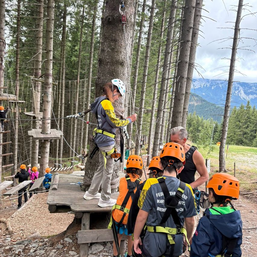 Eine Gruppe von Menschen in Sicherheitsausrüstung versammelt sich um einen Baum im Wald. Eine Frau steht auf einer Holzplattform und gibt möglicherweise Anweisungen. Die Kinder tragen orangefarbene Helme und Rucksäcke. Das Gebiet ist von Bäumen und einer Holzstruktur umgeben.