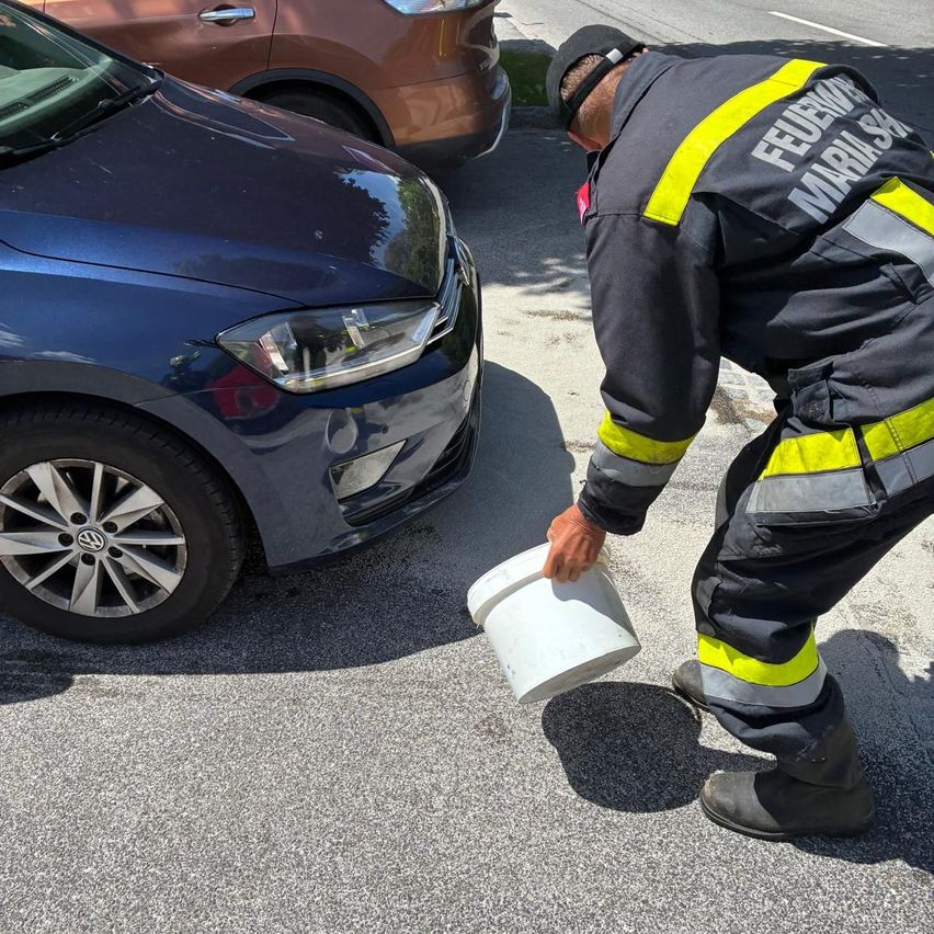 Ein Feuerwehrmann gießt Wasser auf den Boden neben einem blauen Auto mit einem braunen Auto dahinter an einem sonnigen Tag.