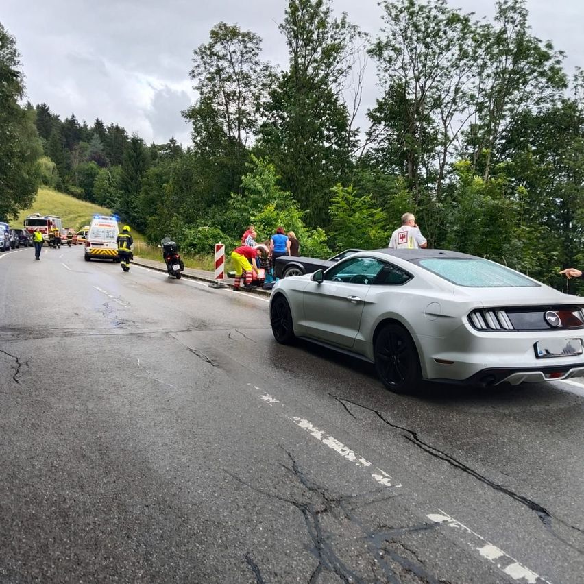 A silver sports car sits on the roadside with an accident scene nearby. Several people are attending to an injured individual, while emergency vehicles and personnel are present.