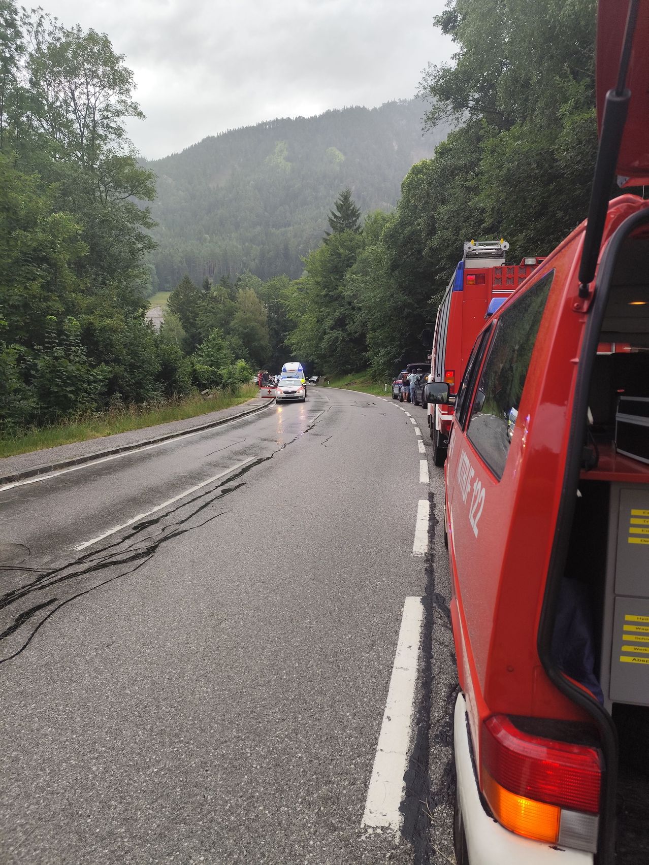 Emergency vehicles are parked on the side of a road. The road has white markings and is surrounded by trees. A person stands near the vehicles.