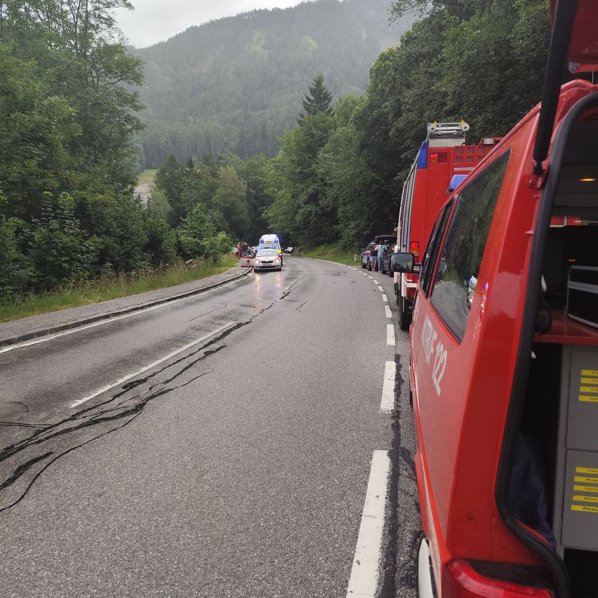 Notfahrzeuge stehen am Straßenrand. Die Straße hat weiße Markierungen und ist von Bäumen umgeben. Eine Person steht in der Nähe der Fahrzeuge.