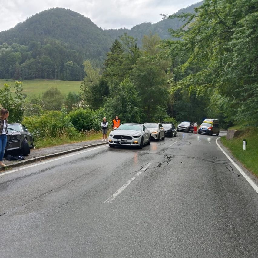 A road with several cars parked on the side. One car is on the road, and a person is standing near the road. There are also trees and mountains in the background.