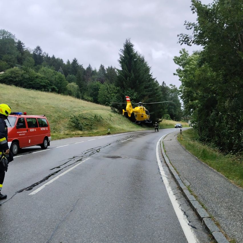An emergency scene on a road. A yellow helicopter hovers above, and a person on a motorcycle is nearby. A red fire truck and a police officer stand by.