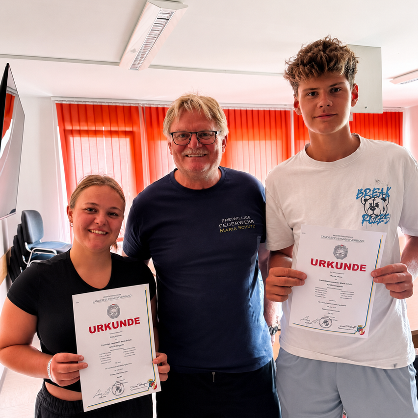 Drei Personen stehen zusammen und halten Urkunden. Der Mann trägt ein marineblaues Shirt, die Frau ein schwarzes Shirt und der Junge ein weißes Shirt mit einem blauen Logo. Sie befinden sich in einem Raum mit orangefarbenen Jalousien und einer weißen Decke.
