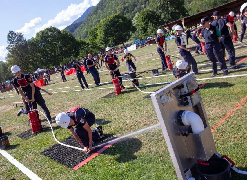 A group of firefighters in uniform are practicing a water relay on a grassy field with spectators in the background.