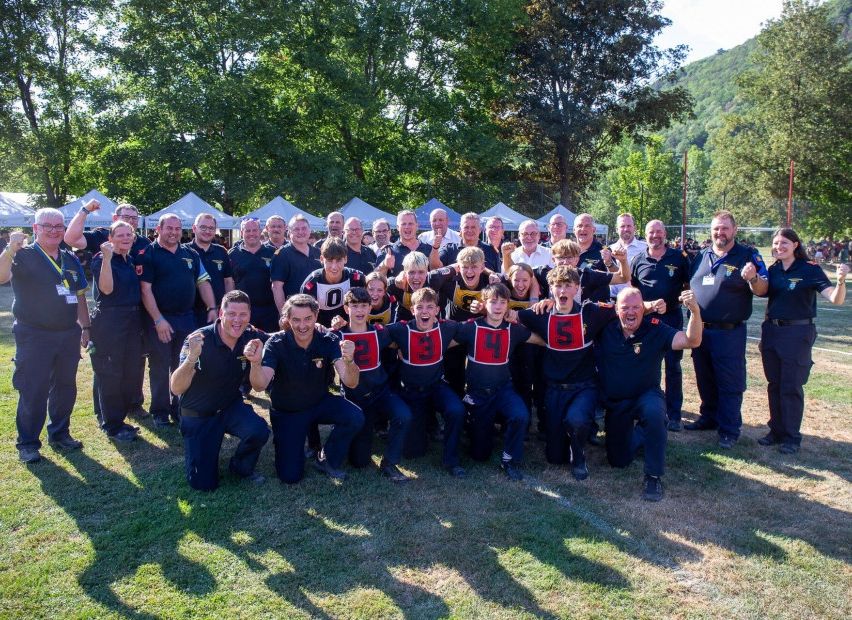 A group of people in matching uniforms pose for a photo on a grassy field with trees and tents in the background.
