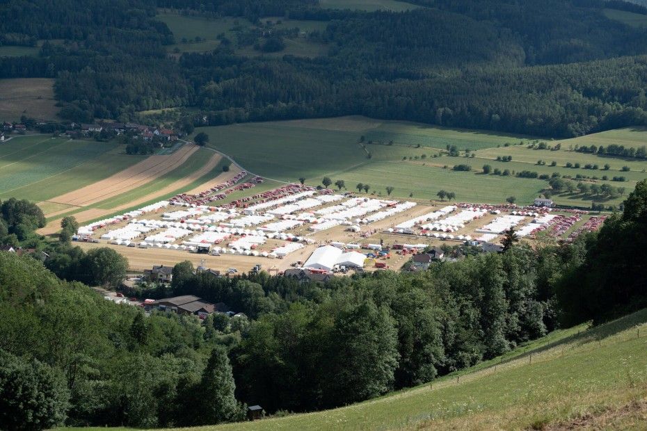 An aerial view of a large outdoor event with numerous white tents set up in rows amidst green fields and dense forest.