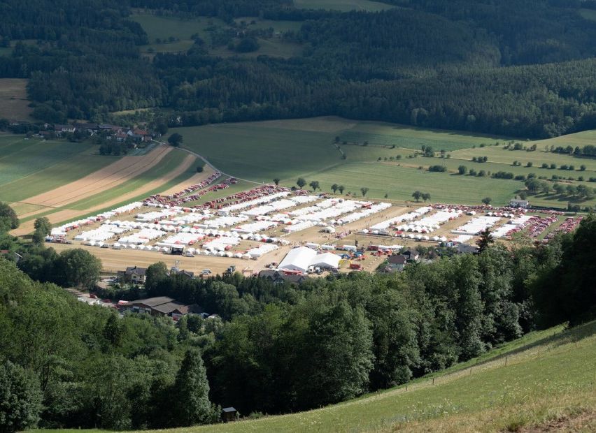 Ein Luftbild von einer großen Freiluftveranstaltung mit zahlreichen weißen Zelten in Reihen, umgeben von grünen Feldern und dichtem Wald.