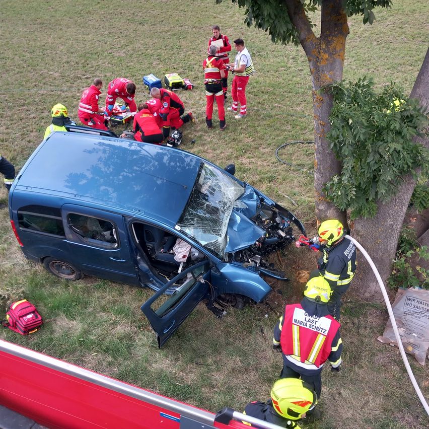 Rettungskräfte kümmern sich um einen blauen Van, der mit einem Baum kollidiert ist. Die Front des Vans ist schwer beschädigt, die Windschutzscheibe ist zersplittert und die Motorhaube ist eingedrückt. Mehrere Personen in roten Uniformen kümmern sich um die Insassen des Fahrzeugs.