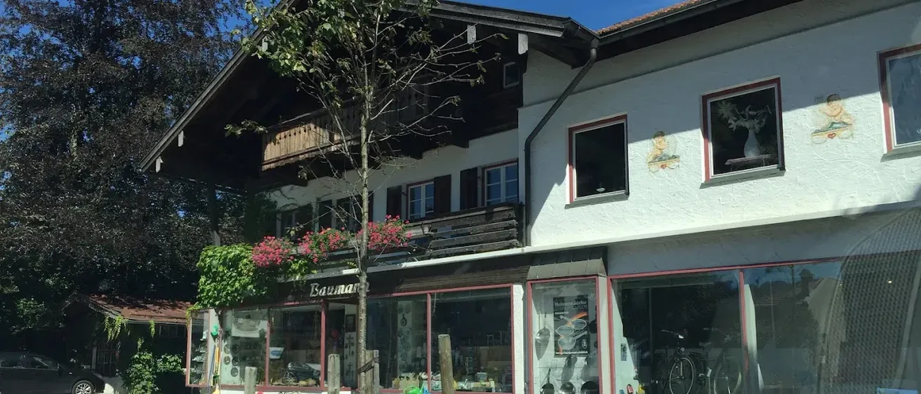 Ein weißes Gebäude mit rotem Rand, das ein Schild mit der Aufschrift Baumarkt zeigt, steht an einer Straße. Es verfügt über einen Balkon mit rosa Blumen und mehrere Fenster. Fahrräder sind im Laden geparkt, und die Umgebung umfasst Bäume und einen Rasenbereich.
