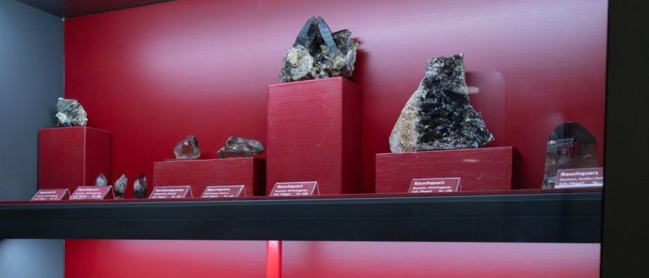 Ein Vitrinenschrank mit roten Wänden und schwarzen Regalen zeigt verschiedene Mineralien, jedes auf einem roten Podest, beschriftet mit Namen wie Rauchquarz und Amethyst.