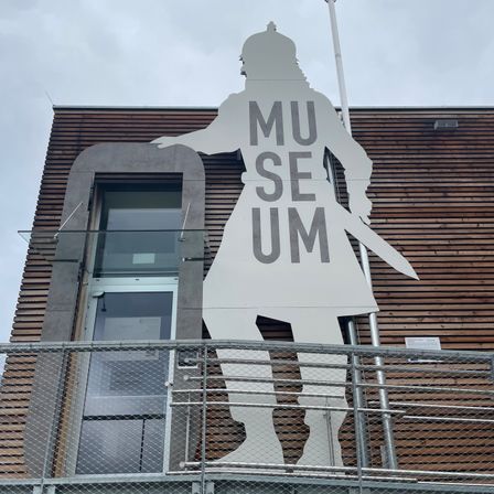 Eine große, weiße Silhouette einer Person, die ein Schwert hält, ist an der Außenwand eines Gebäudes befestigt. Über der Figur steht 'MU' und darunter 'SE UM'. Das Gebäude hat eine Holzwand und ein Glasfenster.