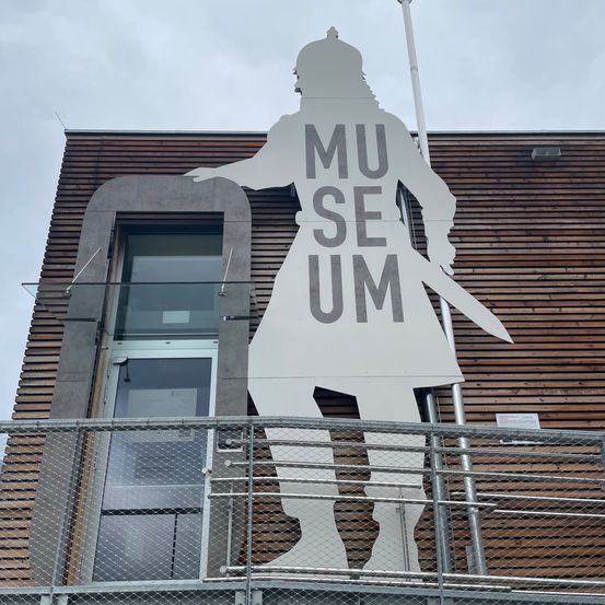 Eine große, weiße Silhouette einer Person, die ein Schwert hält, ist an der Außenwand eines Gebäudes befestigt. Über der Figur steht 'MU' und darunter 'SE UM'. Das Gebäude hat eine Holzwand und ein Glasfenster.