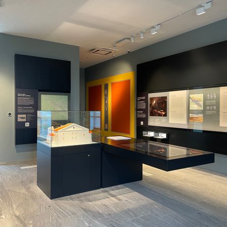 Eine moderne Galerie mit einer Glasvitrine, die ein kleines Gebäudemodell zeigt. Der Raum hat eine graue Wand und Boden, mit mehreren schwarzen und gelben Paneelen an der Wand. Es gibt mehrere Lichter an der Decke.