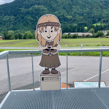 Eine Ausschnitt der lächelnden Mädchen mit einem Gürtel und einem Stirnband steht auf einer Plattform mit einem Metallzaun. Dahinter ist ein breites Feld mit Bergen in der Ferne.