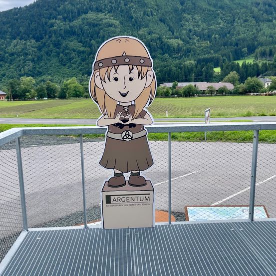 Eine Ausschnitt der lächelnden Mädchen mit einem Gürtel und einem Stirnband steht auf einer Plattform mit einem Metallzaun. Dahinter ist ein breites Feld mit Bergen in der Ferne.