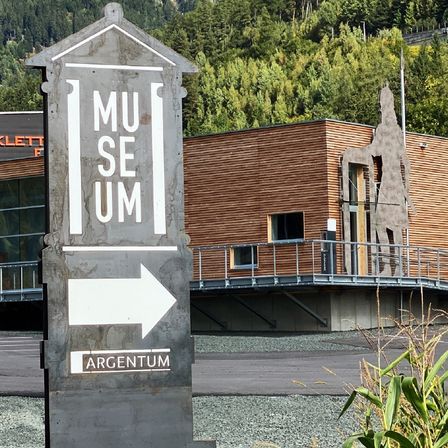 Ein grauer Richtungspfeil weist nach rechts und zeigt den Begriff Argentum. Dahinter befindet sich ein modernes Gebäude mit einer Statue einer Person auf dem Balkon.