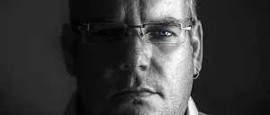 Ein Mann mit Brille schaut direkt in die Kamera, seine Augen leicht geöffnet, mit einem ernsten Ausdruck auf dunklem Hintergrund.