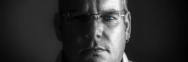 Ein Mann mit Brille schaut direkt in die Kamera, seine Augen leicht geöffnet, mit einem ernsten Ausdruck auf dunklem Hintergrund.