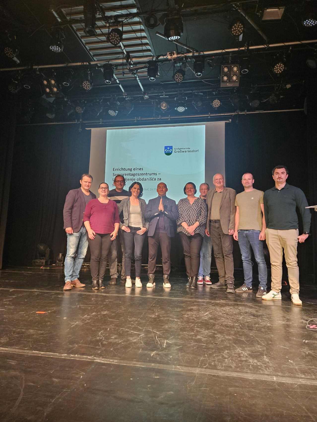 Eine Gruppe von Menschen steht auf einer Bühne mit einer Projektionsleinwand hinter ihnen, die Informationen über ein Seniorenzentrum anzeigt. Sie präsentieren oder feiern wahrscheinlich etwas.