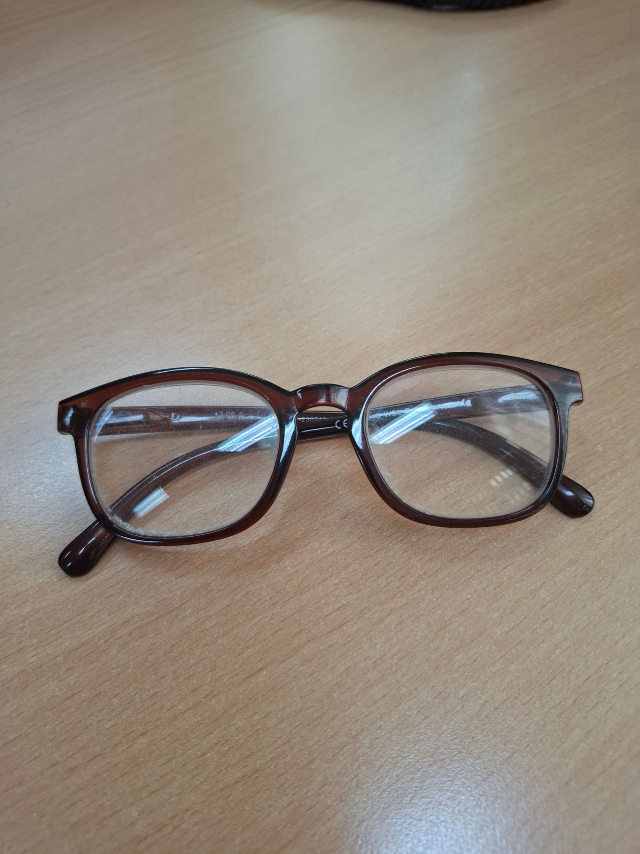 Ein Brillengestell mit braunen Bügeln und klaren Gläsern liegt auf einer Holzoberfläche. Die Gläser sehen sauber aus.