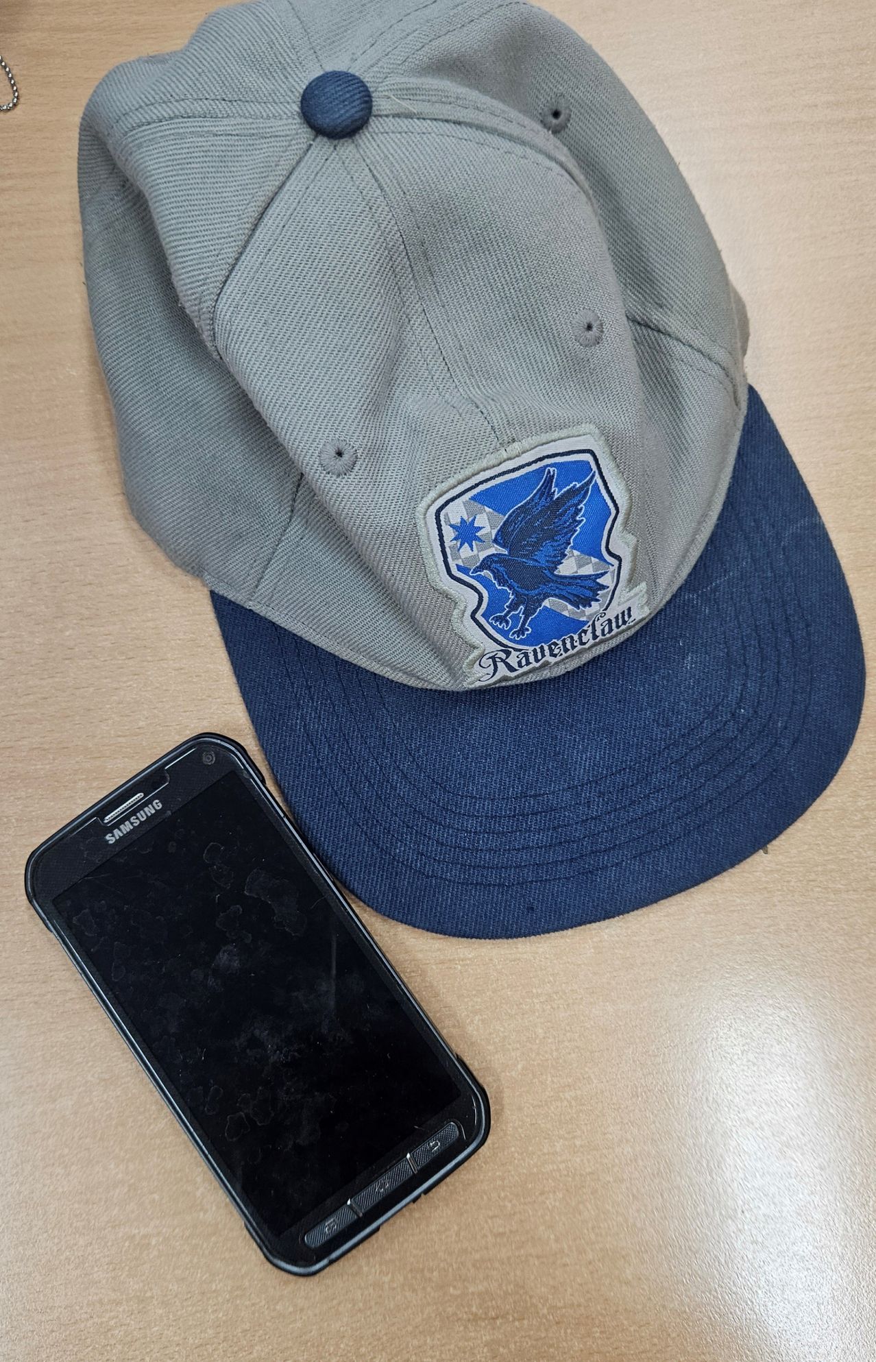 Eine graue und blaue Baseballkappe mit einem blau-weißen Logo eines Vogels und dem Wort 'Ravenclaw' darauf, neben einem schwarzen Samsung-Smartphone.