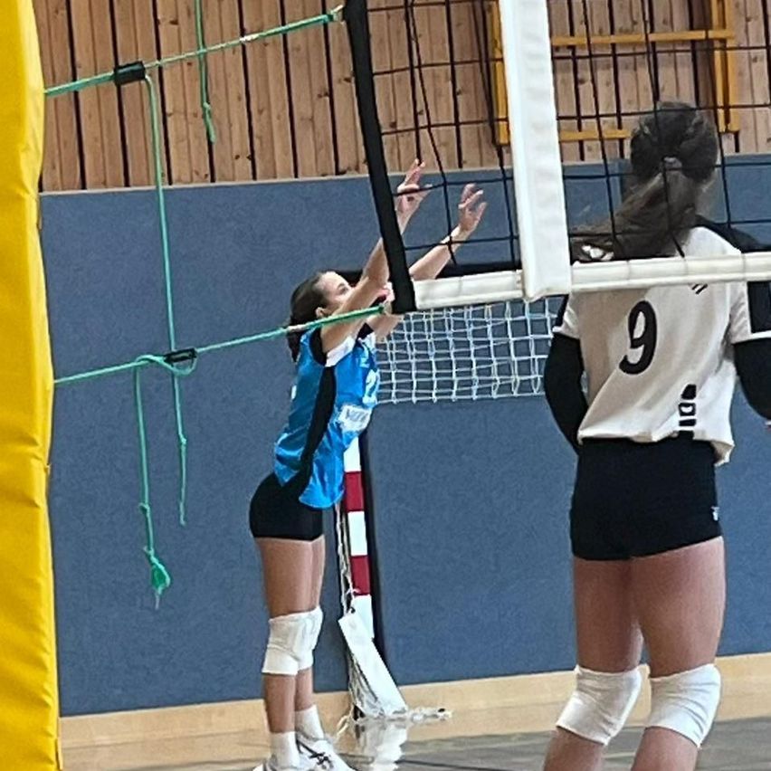Zwei Frauen spielen Volleyball. Eine springt, um den Ball zu schlagen, und die andere beobachtet. Beide tragen Sportbekleidung. Der Platz hat ein Netz und eine gelbe Barriere.