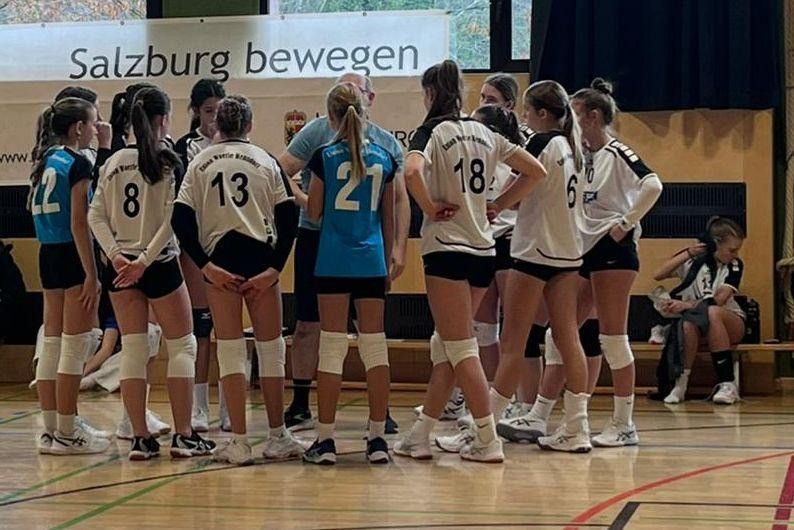Eine Volleyballmannschaft ist auf einem Spielfeld versammelt und hört einem Mann in einem blauen Shirt zu. Sie tragen nummerierte Trikots und Knieschoner. Ein Banner steht für 'Salzburg bewegen'.
