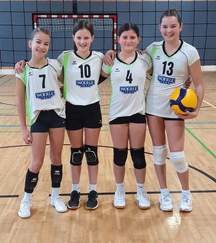 Vier Mädchen in weißen Trikots mit den Nummern 7, 10, 4 und 13 stehen lächelnd auf einem Volleyballfeld. Sie tragen Knieschoner und Turnschuhe. Ein Netz ist im Hintergrund.