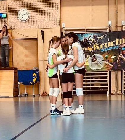 Eine Gruppe junger Frauen in Volleyball-Uniformen umarmt sich in einer Turnhalle. Eine Frau steht hinter ihnen auf einer Bühne, und ein Banner ist an der Wand angebracht.