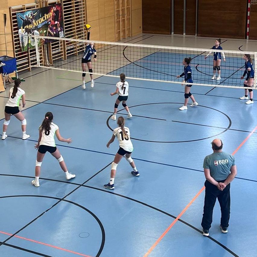 Eine Gruppe von weiblichen Volleyballspielerinnen in blauen Uniformen spielt auf einem Hallenplatz mit Netz. Eine Spielerin springt, um den Ball zu schlagen.