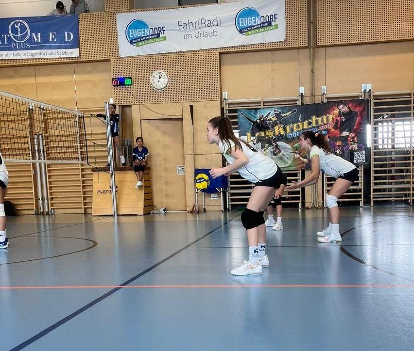 Eine Gruppe junger Mädchen spielt Volleyball auf einem Hallenplatz. Eines der Mädchen hält einen Ball, während andere bereit sind, ihn zu schlagen. Eine Erwachsene steht in der Nähe des Netzes. An der Wand hängen Banner, darunter eine Uhr.