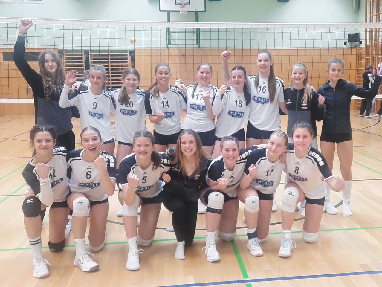 Eine Gruppe weiblicher Volleyballspielerinnen in Uniform posiert für ein Foto in einer Turnhalle. Einige Spielerinnen haben Nummern auf ihren Trikots. Eine Frau in Schwarz steht in der Mitte.