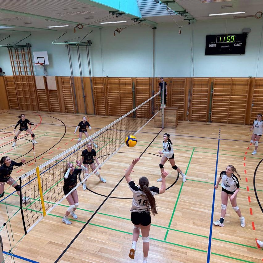 Eine Gruppe von weiblichen Volleyballspielerinnen spielt in einer Turnhalle. Eine Spielerin springt, um den Ball über das Netz zu schlagen. Der Platz hat blaue und grüne Markierungen. Eine Anzeigetafel ist an der Wand angebracht.