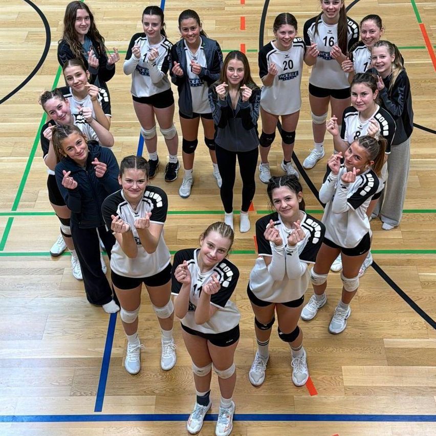 Eine Volleyballmannschaft posiert für ein Foto auf einem Spielfeld und trägt weiße Trikots mit Nummern und schwarze Shorts. Einige Spieler tragen Knieschoner. Das Spielfeld hat blaue und grüne Markierungen.