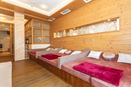 Ein holzverkleideter Raum mit mehreren Lounge-Betten. Die Betten sind mit weichen Kissen und burgunderroten Decken ausgestattet. Holzbalken säumen die Wände.