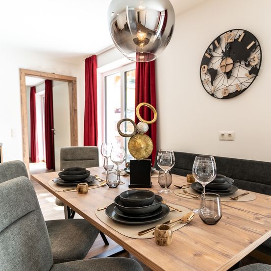 Ein Essbereich mit einem Holztisch, schwarzen Tellern und einer dekorativen Uhr an der Wand. Der Raum hat ein modernes Design mit roten Vorhängen und einer silbernen Lampe.