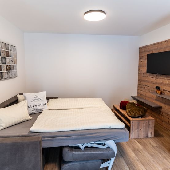Ein Schlafzimmer mit einem Bett, Kissen und einem Fernseher an einer Holzwand. Das Zimmer hat eine graue Couch, einen Holztisch und eine Deckenlampe.