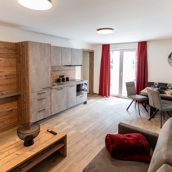 Ein offenes Wohnzimmer mit einer Couch und einem Couchtisch. Die Küche ist mit Schränken, einem Waschbecken und einem Ofen ausgestattet. Ein Esstisch und Stühle befinden sich in der Nähe der Tür.