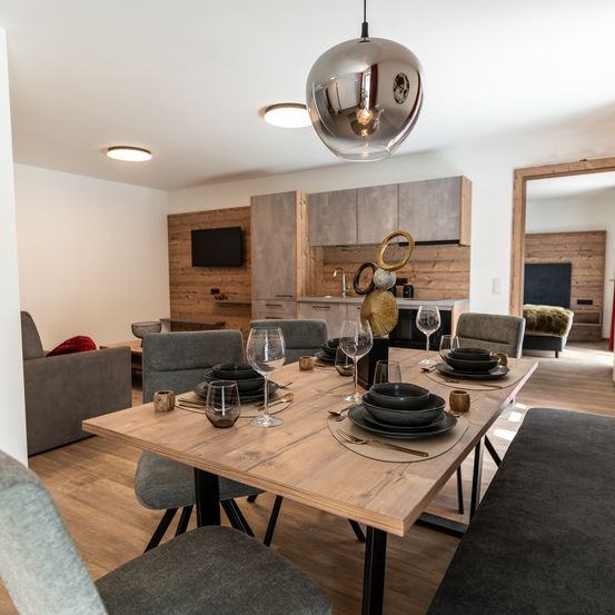 Ein Essbereich mit einem hölzernen Tisch, der für das Abendessen gedeckt ist. Eine moderne Pendelleuchte hängt von der Decke. Im Hintergrund befindet sich eine Küche mit Holzwänden und einem an der Wand montierten Fernseher.