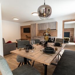 Ein Essbereich mit einem hölzernen Tisch, der für das Abendessen gedeckt ist. Eine moderne Pendelleuchte hängt von der Decke. Im Hintergrund befindet sich eine Küche mit Holzwänden und einem an der Wand montierten Fernseher.