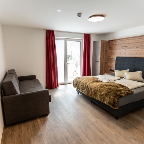 Ein gemütliches Schlafzimmer mit Holzboden, einem Bett, das mit einer braunen Pelzdecke bedeckt ist, einem Sofa und einer Glastür mit roten Vorhängen.