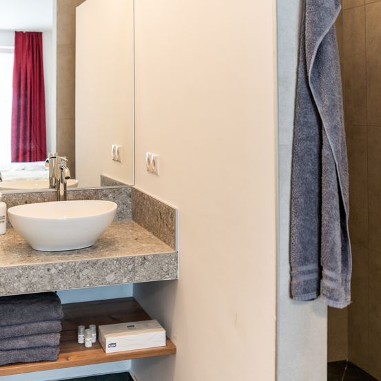 Ein Badezimmer mit Marmorarbeitsplatte zeigt ein weißes Waschbecken und einen Spiegel, der einen roten Vorhang reflektiert. Handtücher und Pflegeprodukte sind ordentlich auf einem Holzregal darunter angeordnet.